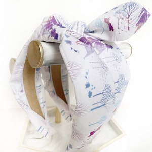 May include: A white headband with a blue and purple watercolor print of a winter scene. The headband is tied in a bow and is displayed on a wooden stand.