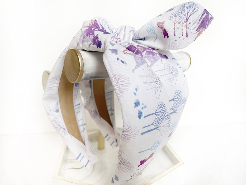 May include: A white headband with a blue and purple watercolor print of a winter scene. The headband is tied in a bow and is displayed on a wooden stand.