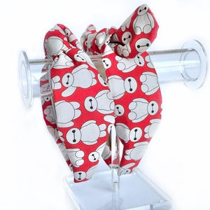 May include: Two red hair scrunchies with a white pattern of cartoon robots. The scrunchies are tied together in a bow.