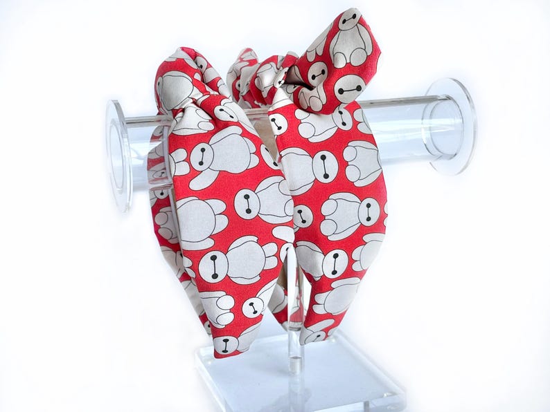 May include: Two red hair scrunchies with a white pattern of cartoon robots. The scrunchies are tied together in a bow.