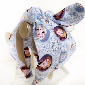 May include: A blue headband with a white background and a pattern of snowflakes and images of Elsa and Anna from the movie Frozen. The headband has a bow on top and the text "Mythic Journey" is printed on the headband.