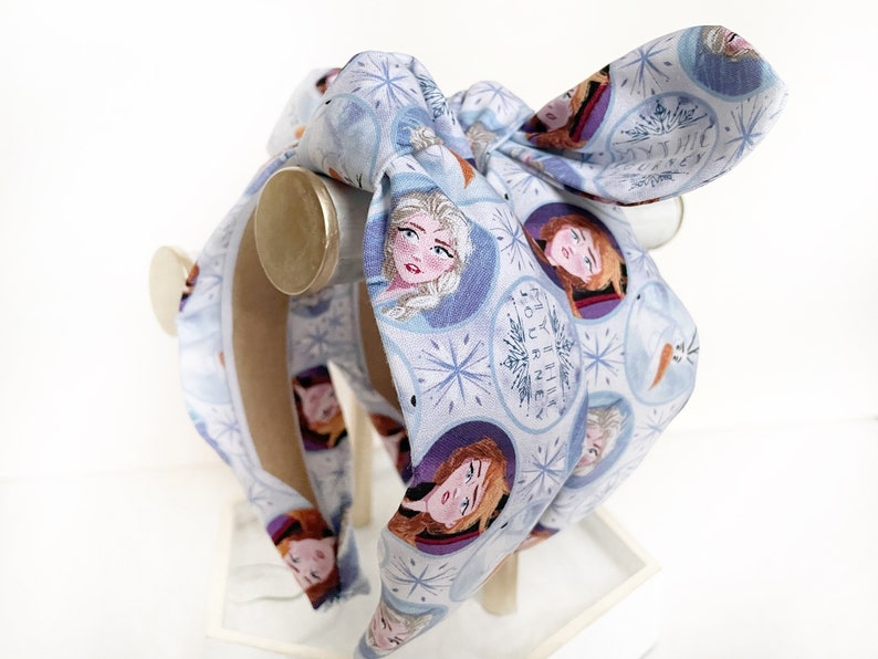 May include: A blue headband with a white background and a pattern of snowflakes and images of Elsa and Anna from the movie Frozen. The headband has a bow on top and the text "Mythic Journey" is printed on the headband.