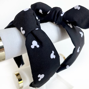 K&ouml;nnte beinhalten: Schwarzes Stirnband mit einem wei&szlig;en Mickey-Maus-Muster. Das Stirnband ist oben zu einer Schleife gebunden.