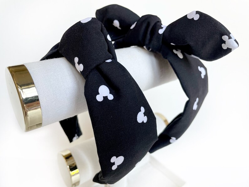 K&ouml;nnte beinhalten: Schwarzes Stirnband mit einem wei&szlig;en Mickey-Maus-Muster. Das Stirnband ist oben zu einer Schleife gebunden.