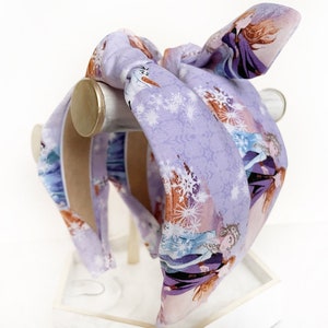 May include: Purple headband with a snowflake pattern and images of the characters from the movie Frozen. The headband is tied in a bow at the top.