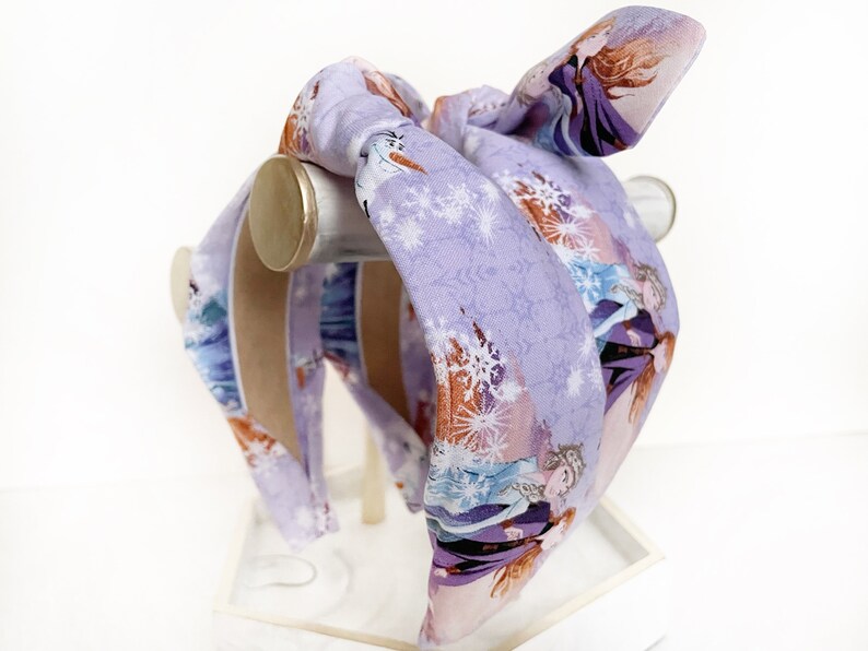 May include: Purple headband with a snowflake pattern and images of the characters from the movie Frozen. The headband is tied in a bow at the top.