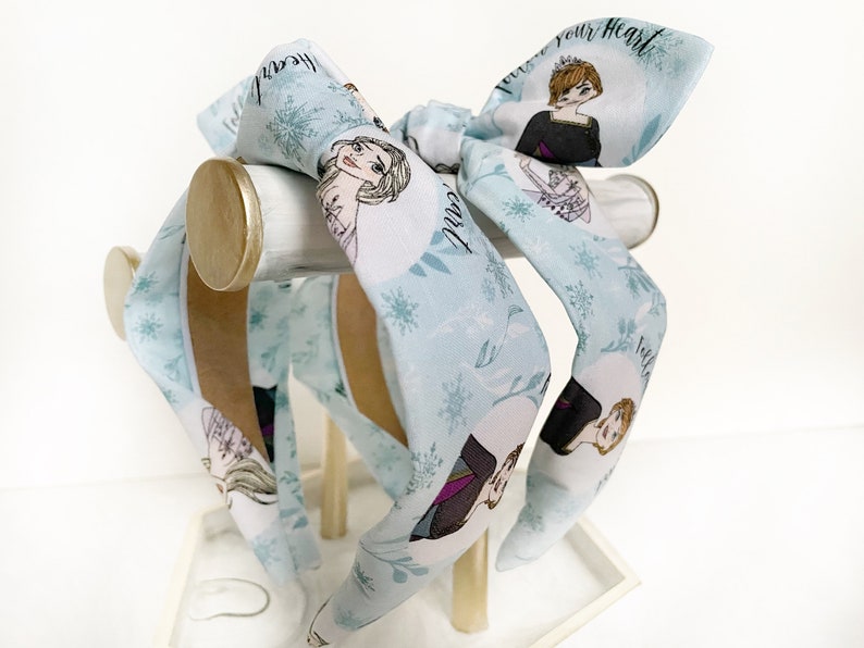 May include: Three blue headbands with a white background and a snowflake pattern. The headbands feature images of the characters from the movie Frozen, including Elsa and Anna. The text "Let it go" and "Follow your heart" are printed on the headbands.