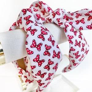 K&ouml;nnte beinhalten: Ein wei&szlig;es Stirnband mit einem roten Polka-Dot-Schleifenmuster. Das Stirnband ist oben zu einem Knoten gebunden.
