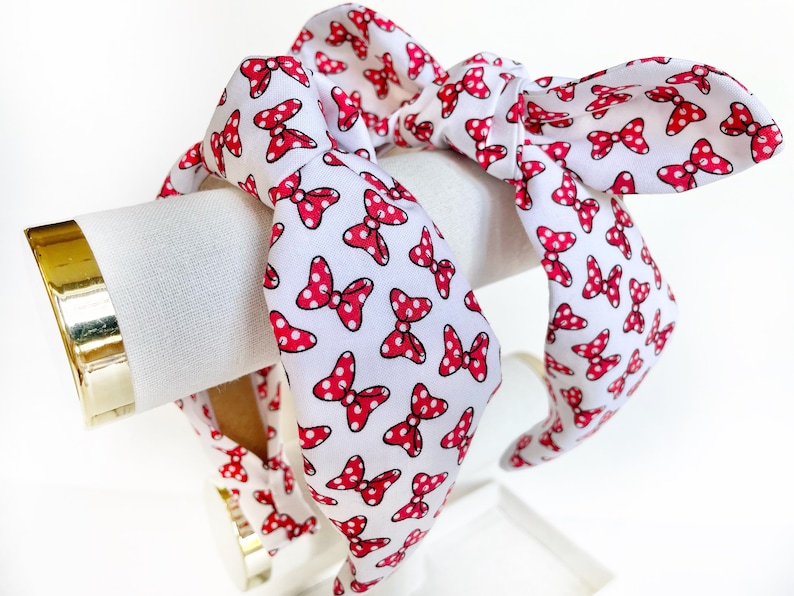 K&ouml;nnte beinhalten: Ein wei&szlig;es Stirnband mit einem roten Polka-Dot-Schleifenmuster. Das Stirnband ist oben zu einem Knoten gebunden.