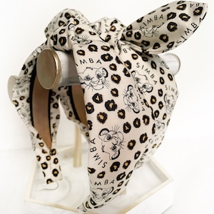 May include: A cream-colored headband with a bow, featuring a repeating pattern of Simba from The Lion King, black leopard spots, and the name "Simba" in black. The headband is displayed on a white stand.