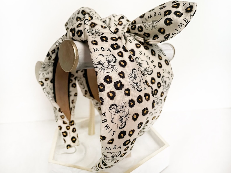 May include: A cream-colored headband with a bow, featuring a repeating pattern of Simba from The Lion King, black leopard spots, and the name "Simba" in black. The headband is displayed on a white stand.