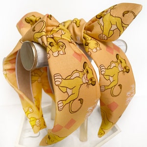 May include: A tan headband with a bow, featuring a repeating pattern of a cartoon lion cub. The headband has a wide band and a bow at the top. The lion cub is yellow and brown, with a smiling expression. The background has a diamond pattern.
