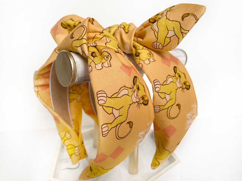 May include: A tan headband with a bow, featuring a repeating pattern of a cartoon lion cub. The headband has a wide band and a bow at the top. The lion cub is yellow and brown, with a smiling expression. The background has a diamond pattern.