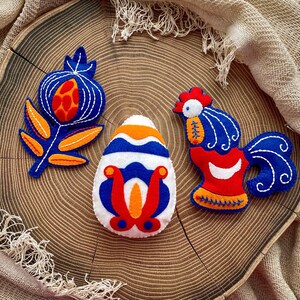 May include: Three felt Easter decorations on a wood slice. A blue and orange floral design, a white egg with blue and orange accents, and a red, white, and blue rooster with white stitching.