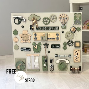 Peut inclure: Un tableau d'activités en bois beige avec divers éléments interactifs pour les tout-petits. Comprend des engrenages, une horloge, un téléphone, une fermeture éclair et un hochet en forme d'ours. Le tableau comporte un hélicoptère, une montgolfière et les chiffres 1 à 9. Le texte "FREE STAND" est visible.