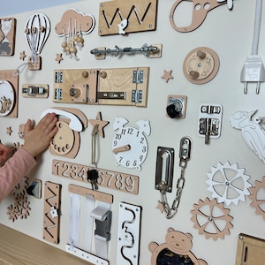 Beige busy board with a variety of interactive elements for toddlers to explore, including latches, locks, buttons, and zippers.