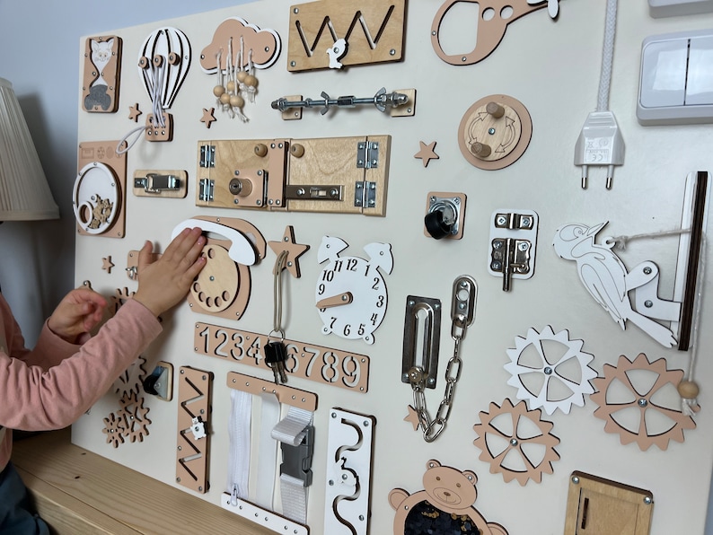 Beige busy board with a variety of interactive elements for toddlers to explore, including latches, locks, buttons, and zippers.