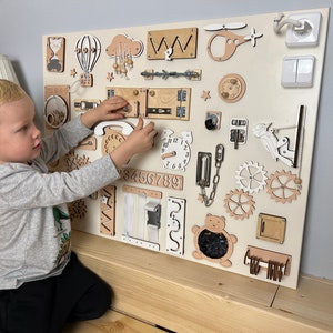 Beige busy board with a variety of interactive elements for toddlers to explore, including latches, locks, buttons, and zippers.