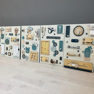 May include: Four light-coloured wooden activity boards with interactive elements. Each board features gears, switches, numbers, and tactile components in blue, beige, and natural wood tones, designed for sensory exploration.
