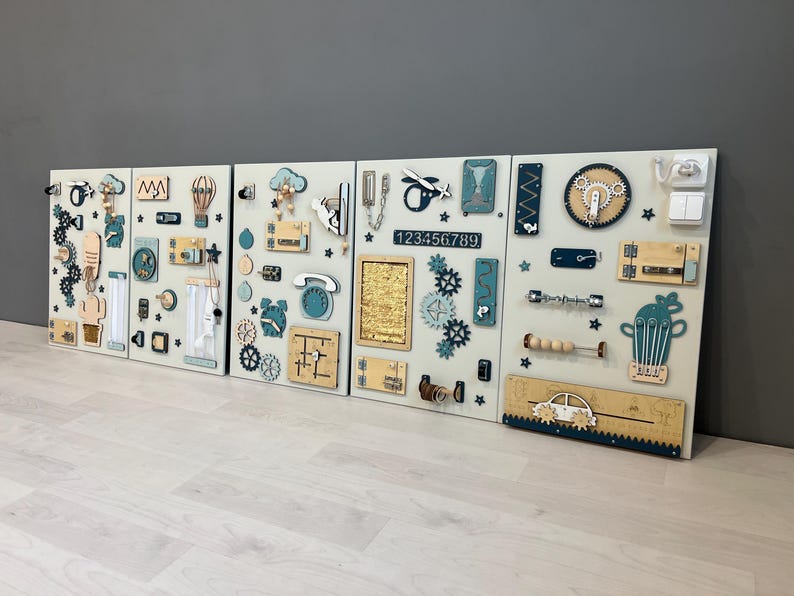 May include: Four light-coloured wooden activity boards with interactive elements. Each board features gears, switches, numbers, and tactile components in blue, beige, and natural wood tones, designed for sensory exploration.