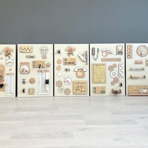 May include: Five wooden activity boards with interactive elements. Each board has a cream background with wooden components such as gears, latches, and shapes. The boards are designed for sensory exploration and learning, with a variety of textures.