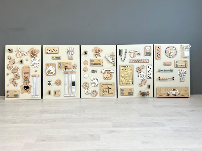 May include: Five wooden activity boards with interactive elements. Each board has a cream background with wooden components such as gears, latches, and shapes. The boards are designed for sensory exploration and learning, with a variety of textures.