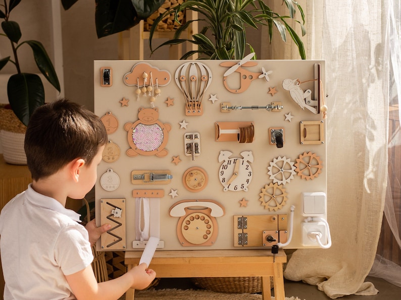 Beige busy board with a variety of interactive elements for toddlers to explore, including latches, locks, buttons, and zippers.