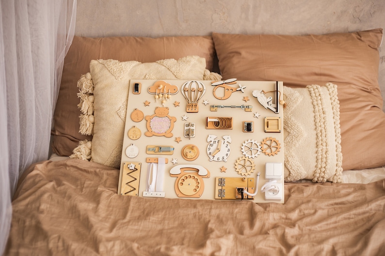 Beige busy board with a variety of interactive elements for toddlers to explore, including latches, locks, buttons, and zippers.