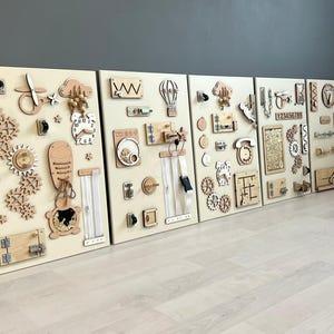 May include: Five wooden activity boards with interactive elements. Each board has different shapes, gears, latches, zippers, and tactile components in natural wood tones. Designed for sensory exploration and learning, with metric measurements.