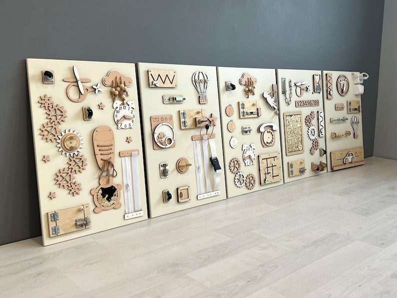 May include: Five wooden activity boards with interactive elements. Each board has different shapes, gears, latches, zippers, and tactile components in natural wood tones. Designed for sensory exploration and learning, with metric measurements.