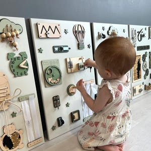 May include: A set of three white wooden activity boards with various interactive elements for toddlers. Features include a clock, zippers, latches, gears, and a shoe with laces. The boards are decorated with green and beige accents.