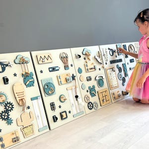 May include: Four white wooden activity boards with interactive elements for children. Includes gears, clocks, zippers, and shapes in blue, beige, and natural wood tones. A young girl in a pink dress interacts with the boards. The boards are made of wood.