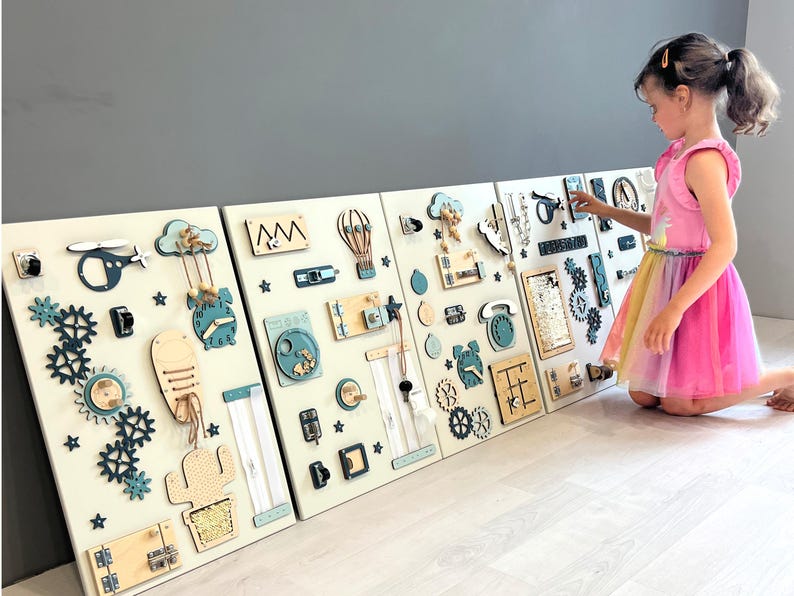 May include: Four white wooden activity boards with interactive elements for children. Includes gears, clocks, zippers, and shapes in blue, beige, and natural wood tones. A young girl in a pink dress interacts with the boards. The boards are made of wood.