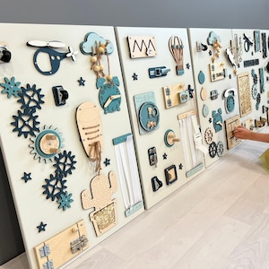 May include: Three white wooden activity boards with interactive elements for children. Includes gears, latches, clocks, a helicopter, and a cactus. The boards are decorated with teal and natural wood accents. The boards are mounted on a wall.