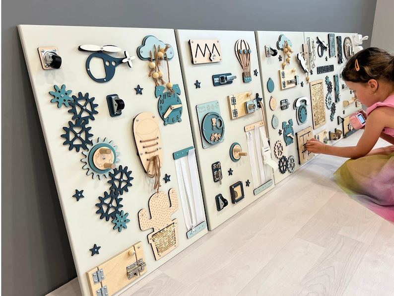 May include: Three white wooden activity boards with interactive elements for children. Includes gears, latches, clocks, a helicopter, and a cactus. The boards are decorated with teal and natural wood accents. The boards are mounted on a wall.
