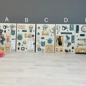May include: Five wooden activity boards with interactive elements. Each board has a light background with gears, latches, and shapes in blue, beige, and natural wood tones. The boards are designed for children's play and learning, with a variety of tactile features.