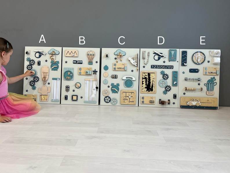 May include: Five wooden activity boards with interactive elements. Each board has a light background with gears, latches, and shapes in blue, beige, and natural wood tones. The boards are designed for children's play and learning, with a variety of tactile features.
