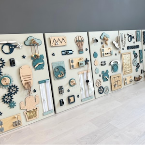 May include: Four wooden activity boards with interactive elements. Each board has a light beige background with attached gears, zippers, clocks, and other shapes in teal, beige, and natural wood tones. The boards are designed to stimulate fine motor skills in children.