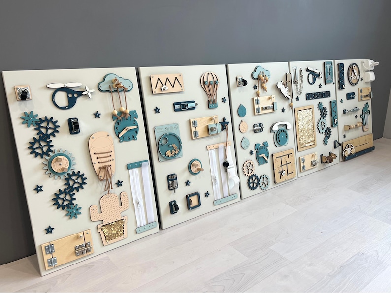 May include: Four wooden activity boards with interactive elements. Each board has a light beige background with attached gears, zippers, clocks, and other shapes in teal, beige, and natural wood tones. The boards are designed to stimulate fine motor skills in children.
