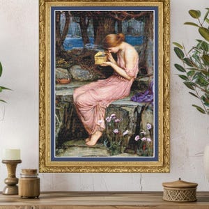 May include: A framed print depicting a woman in a pink dress, seated on a stone wall, looking at a small golden box. The artwork has a forest and river backdrop, and is enclosed in a gold frame with a blue mount.