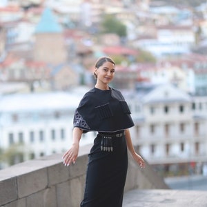 May include: A woman wearing a black dress with a black cape and a silver belt with tassels. The cape has a white embroidered design. She is standing on a stone wall with a city in the background.