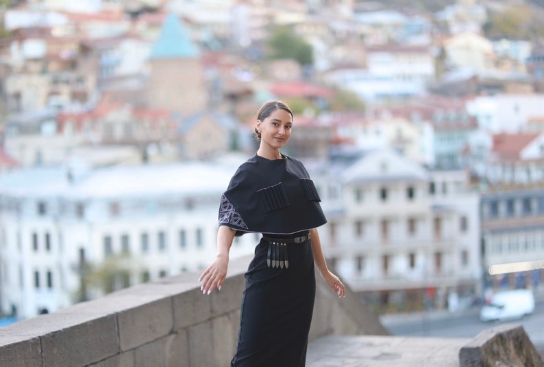 May include: A woman wearing a black dress with a black cape and a silver belt with tassels. The cape has a white embroidered design. She is standing on a stone wall with a city in the background.