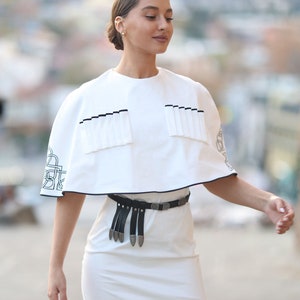 May include: A white dress with a white cape top and a black belt with silver accents. The cape top has a black trim and a pleated detail.