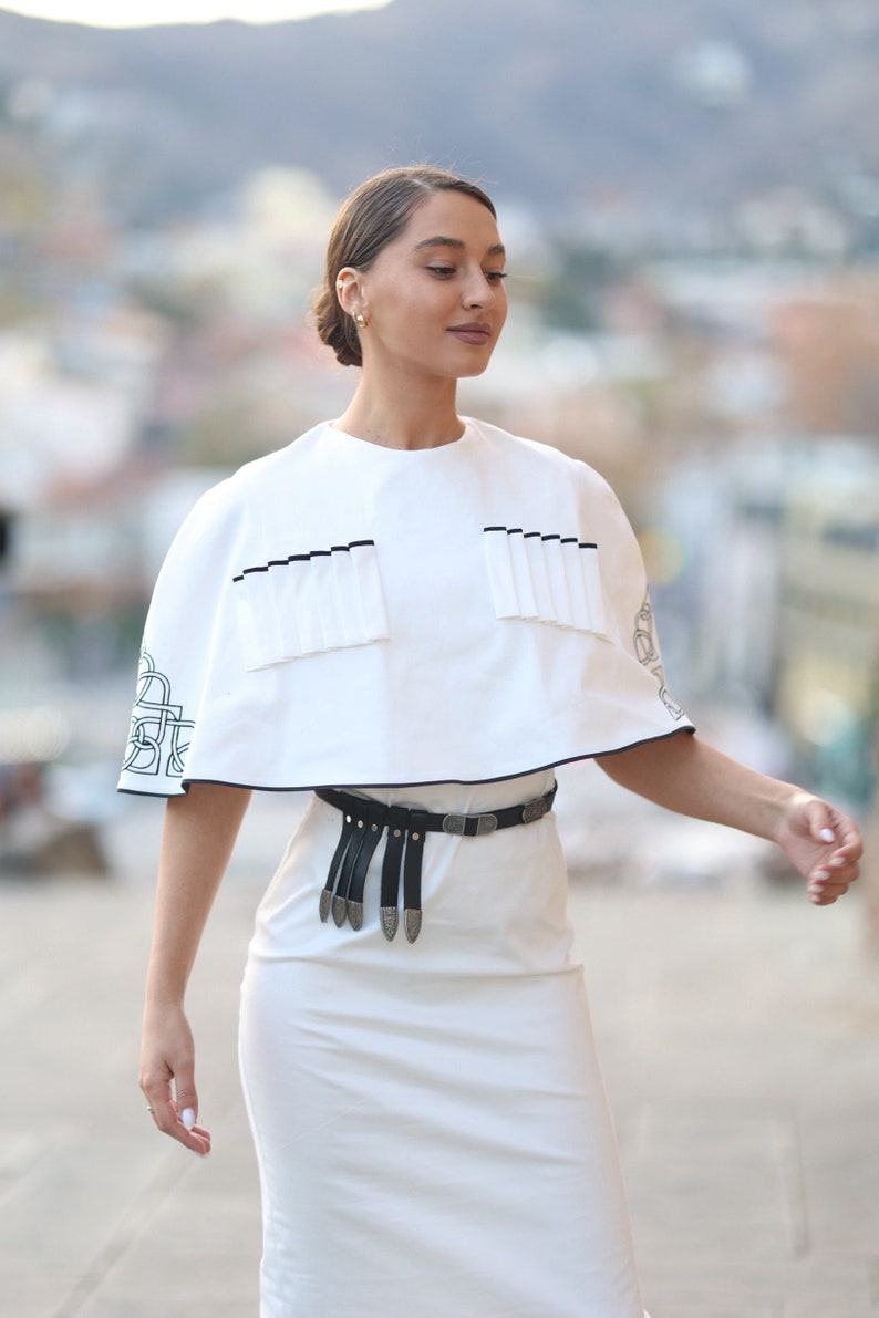 May include: A white dress with a white cape top and a black belt with silver accents. The cape top has a black trim and a pleated detail.