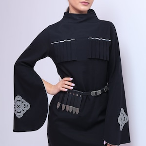 Puede incluir: Un vestido negro de manga larga con ribete blanco. El vestido tiene un cintur&oacute;n decorativo con detalles plateados y un dise&ntilde;o de nudo celta en las mangas.