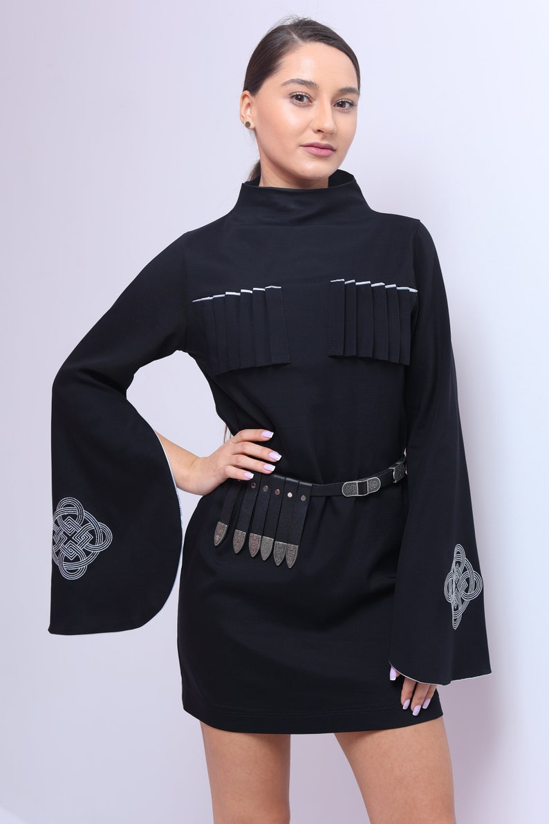 Puede incluir: Un vestido negro de manga larga con ribete blanco. El vestido tiene un cintur&oacute;n decorativo con detalles plateados y un dise&ntilde;o de nudo celta en las mangas.
