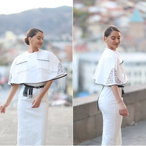 May include: A white dress with a white cape featuring black embroidery. The cape has a black trim and a row of white tassels. The dress has a black belt with silver accents.