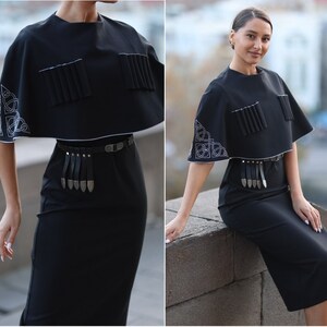 May include: A black dress with a short, black cape-like top. The top has white embroidery and a silver belt with a fringe. The dress is knee-length.