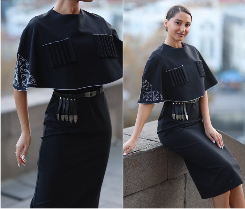 May include: A black dress with a short, black cape-like top. The top has white embroidery and a silver belt with a fringe. The dress is knee-length.