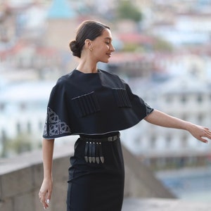 May include: A woman wearing a black dress with a black cape top and a silver belt with a fringe. The cape top has white embroidery on the sleeves.